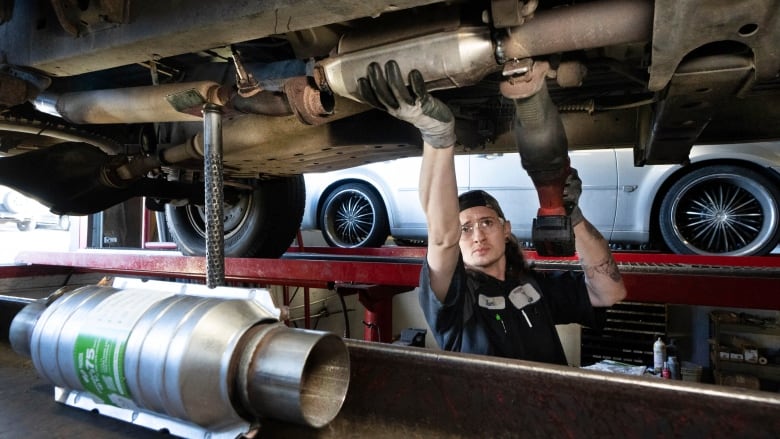 A man stands underneath a car, holding onto a part. In the foreground, a new catalytic converter is seen