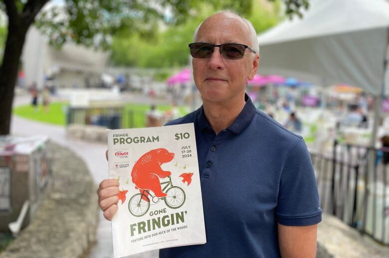 Man wearing a blue shirt stands, holds a pamphlet with the Fringe's programming. 