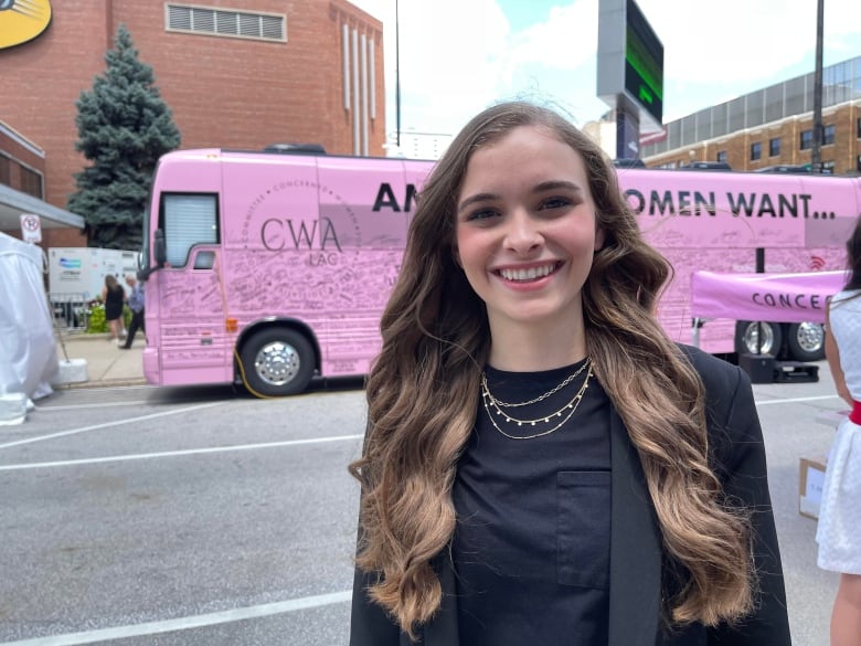 A young woman wearing a black top and gold necklaces poses in front of a pink bus.