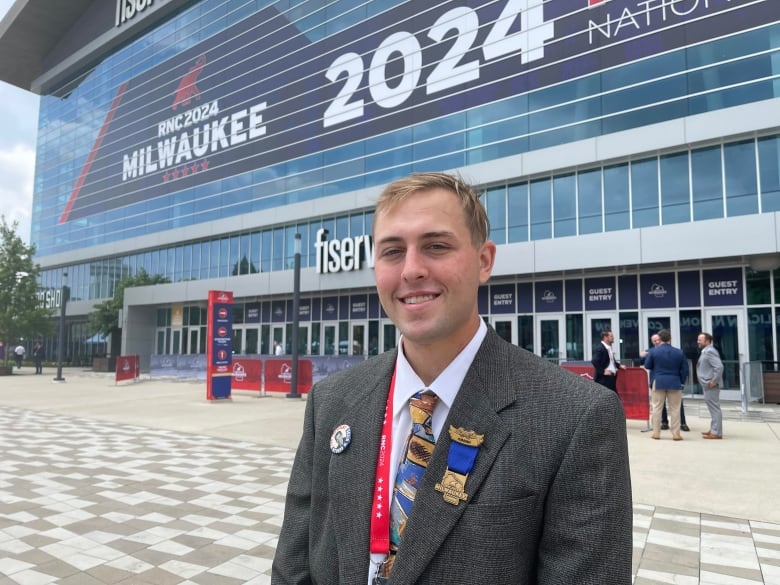 A young man wearing a gray suit jacket with a necktie and buttons on his lapels smiles.