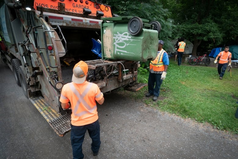 people wearing orange vests dumping trash from a homeless encampment into a dumpster truck