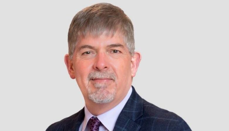 A man with short gray hair, a mustache and goatee, wearing a dark blue checked coat, pale lavender shirt and purple patterned tie looks at the camera for a head and shoulders professional portrait against a white background.