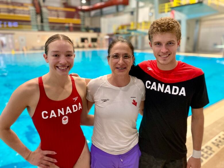 A woman in a red one piece swimsuit, an older woman in a white T-shirt, and a blond, curly-haired man in a Canada shirt link arms while standing in front of a pool. 