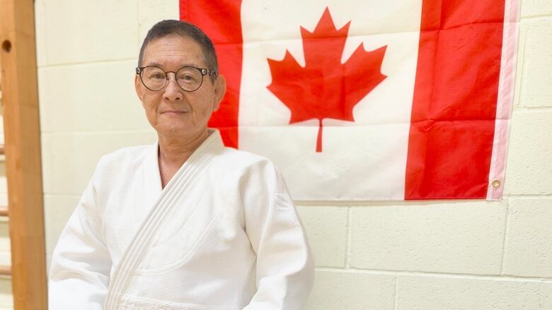 A person in round glasses and a white judo outfit stands in front of a CAnada flag