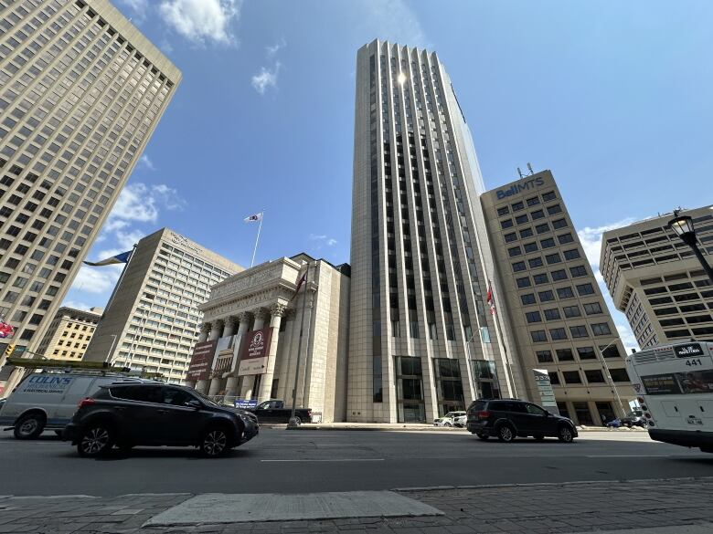 Cars drive through a road in Winnipeg's downtown. Three buildings stand behind the road. 