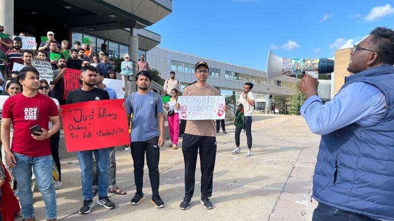 A group of protesters on the left, some of which are holding signs, are watching a man on the right, who is chanting into a megaphone.