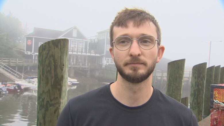 Man in glasses and a black t-shirt stands on a wharf on a foggy day.