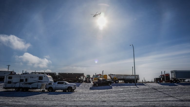 Several trucks and trailers block a snowy highway on a sunny day as a helicopter flies overhead.