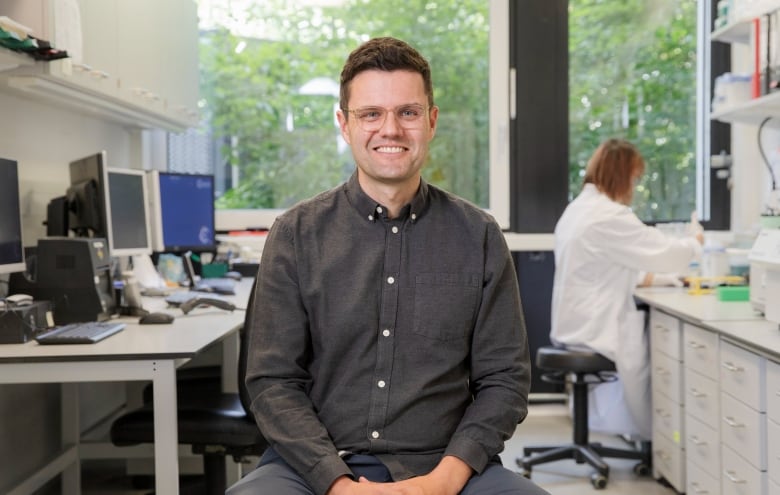 A smiling man with glasses sits beside a row of computers in a lab setting as a woman in a white lab coat works behind him. 