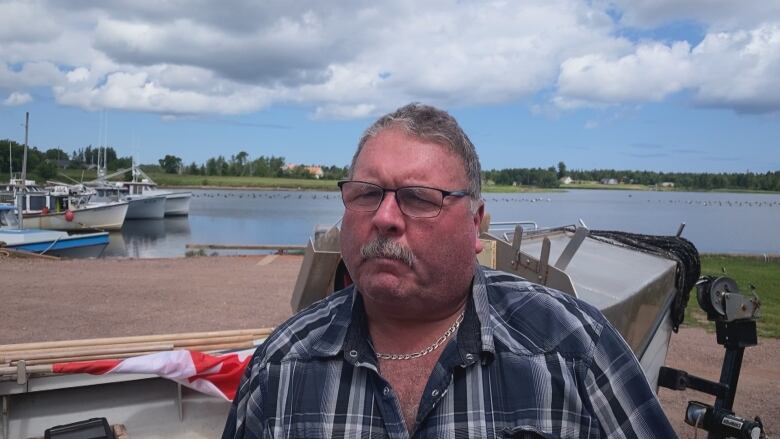 A man with short grey hair, a mustache and glasses speaks in front of a fishing wharf.