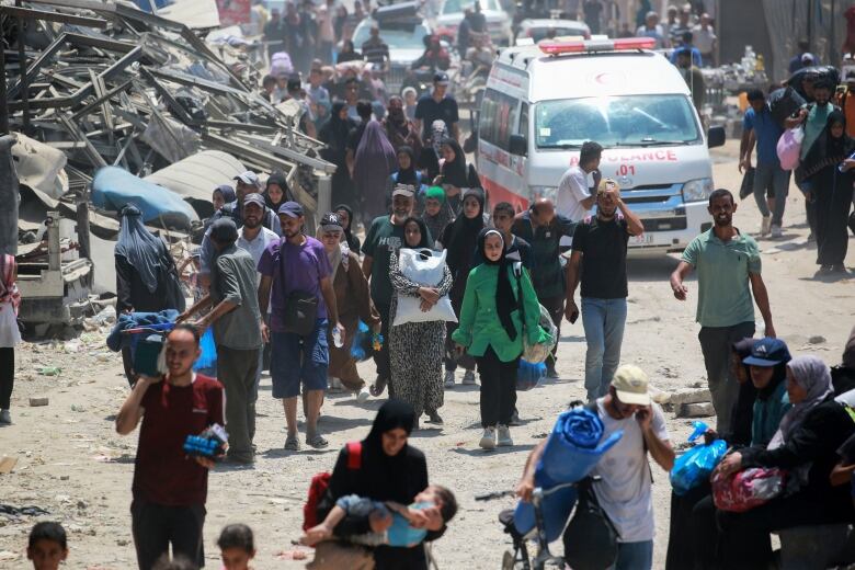 Dozens of people are shown in the photograph walking toward camera on a dirt road, with vehicles and large pieces of metal debris shown.