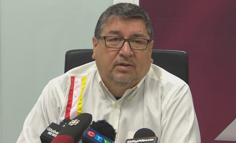 A man with short grey hair sits behind microphones at a table.