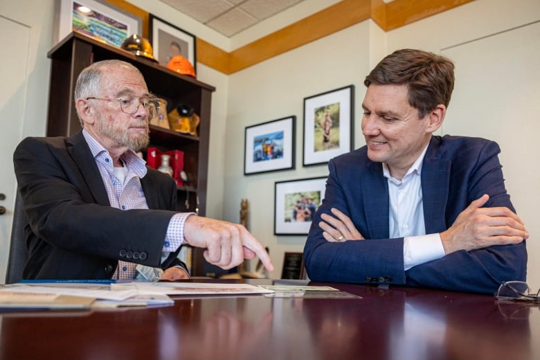 A man points at a dollar bill on a table, sitting next to B.C. Premier David Eby.