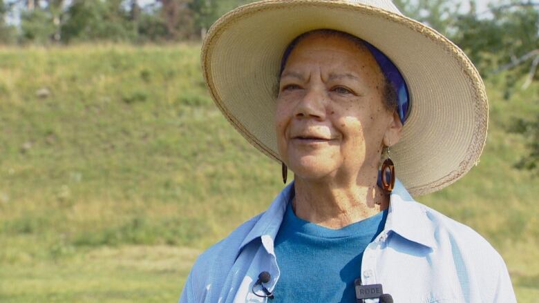 A close-up shot of a woman in large hat. She is wearing a blue T-shirt and a light blue shirt with brown earrings. Grass is visible in the background.