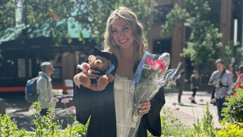 a young woman with blonde hair stands smiling outside. she is holding a bouquet of flowers and a teddy bear. she is wearing a grad gown.