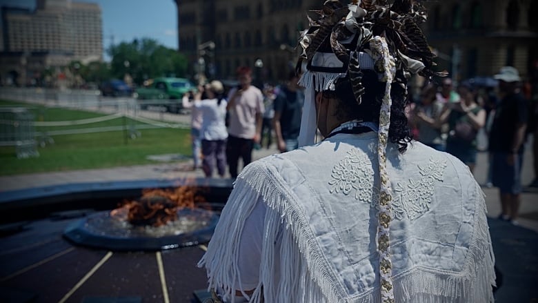 Ramon Kataquapit, a man, stands with his back turned the camera dressed in regalia.