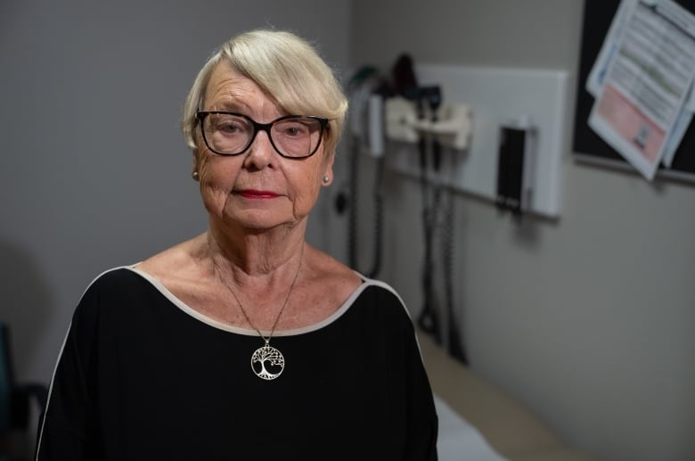 Woman seated in a doctor's office wearing glasses, a black shirt and silver pendant. 