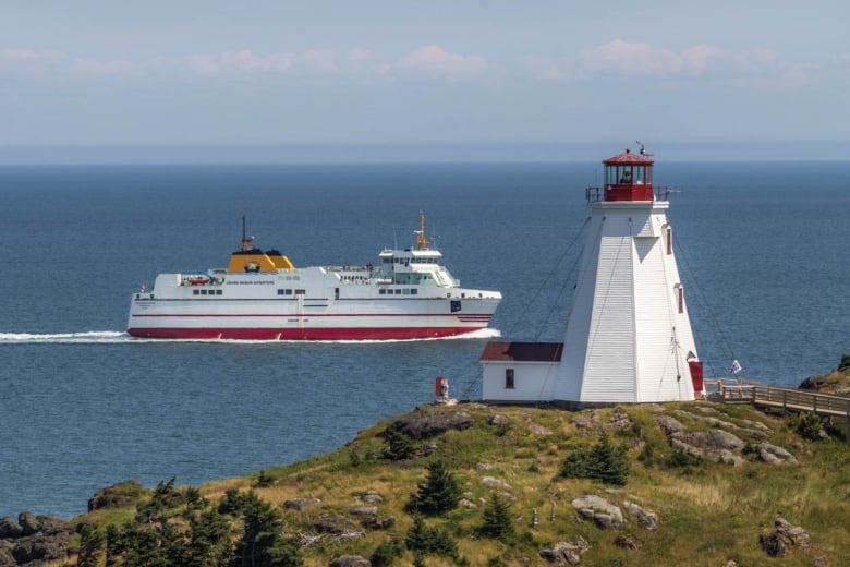 A large white boat moves over blue water in front of a grassy island with a white lighthouse on it.