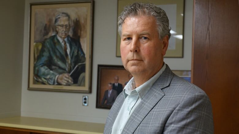 A man with curly grey hair and wearing a grey suit jacket stands in front of a wooden cabinet and a wall with photos and a painting hanging from it.