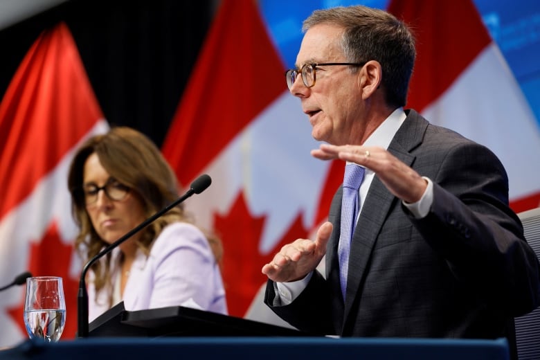 A man gestures while speaking during a news conference and a woman is shown seated to his right.