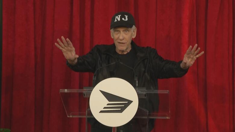 A man in a black outfit and ball cap stands at a podium in front of a curtain.