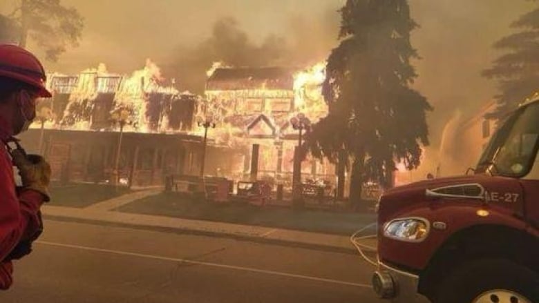 A firefighter looks on as a hotel is aflame. Smoke fills the air. An orange hue fills the scene due to the fire.