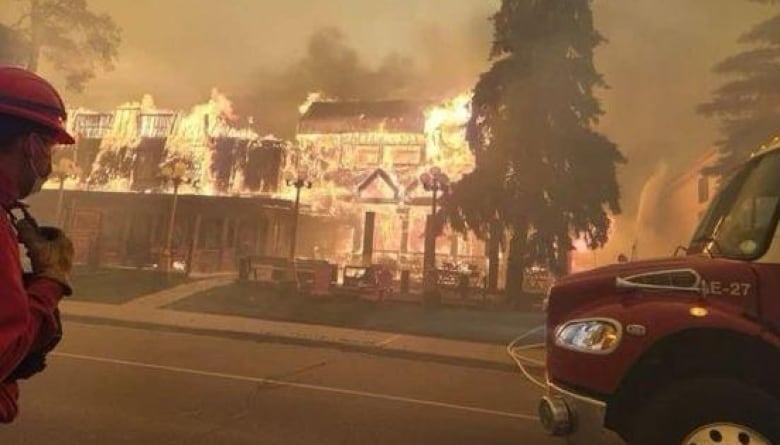 A firefighter looks on as a hotel is aflame. Smoke fills the air. An orange hue fills the scene due to the fire.