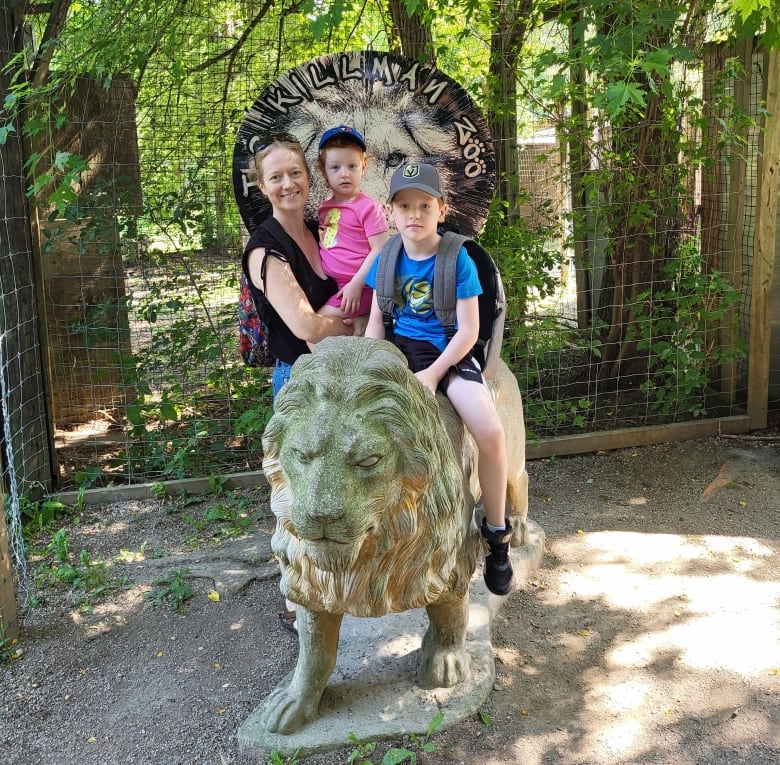 A woman smiles as she holds a young girl next to a boy a few years older who is sitting on a statue of a lion. A sign in the background reads 