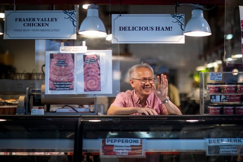 A man stands behind a butcher shop counter, smiling and waving at passersby. 