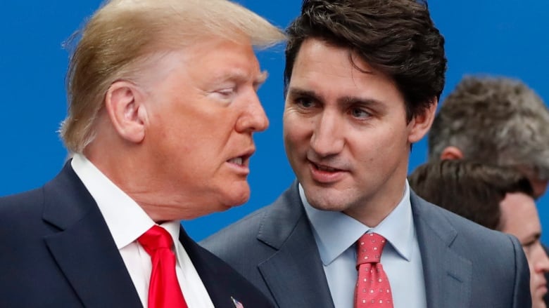 A man with gold hair wearing a navy suit and a red tie speaks while a younger man in a grey suit and red tie looks at him quizzically.