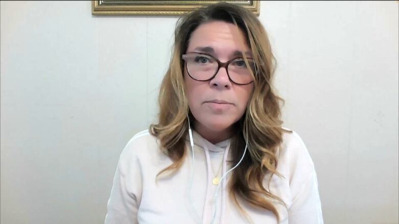 A woman wearing a white sweater sits against a white background. She has earbuds in as she listens to a Zoom call.