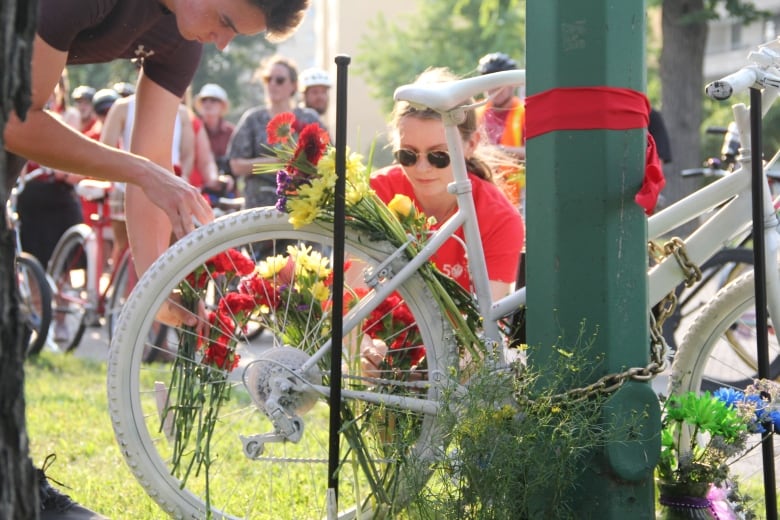 Two people place flowers around the spokes of a bicycle completely painted in white, which was chained to a green pole.