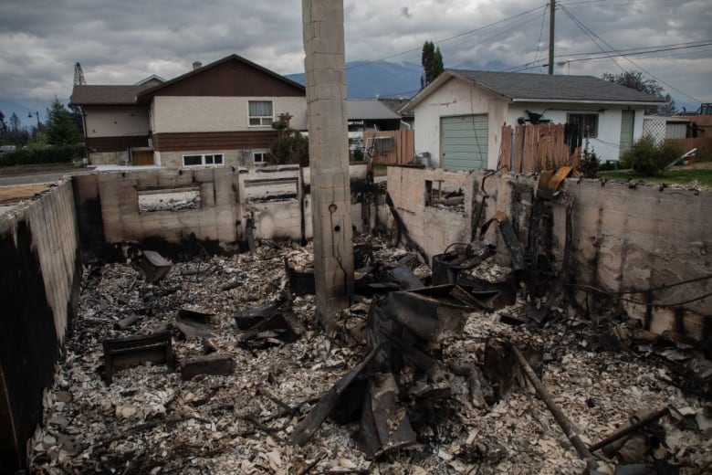 The burned foundation of a home is filled with ash-covered debris and scorched metal. Nearby is a burned wooden fence. Another house seems unharmed.