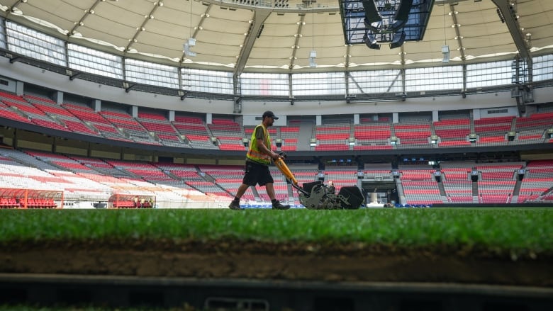 A man in a safety vest pushes a large electric lawnmower across green turf under the dome of a large multi-seat stadium.