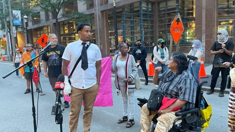 A man in a shirt, tie and cast on his forearm, stands next to microphones at a rally on a city street. An elderly woman in a wheelchair looks at him from his left.