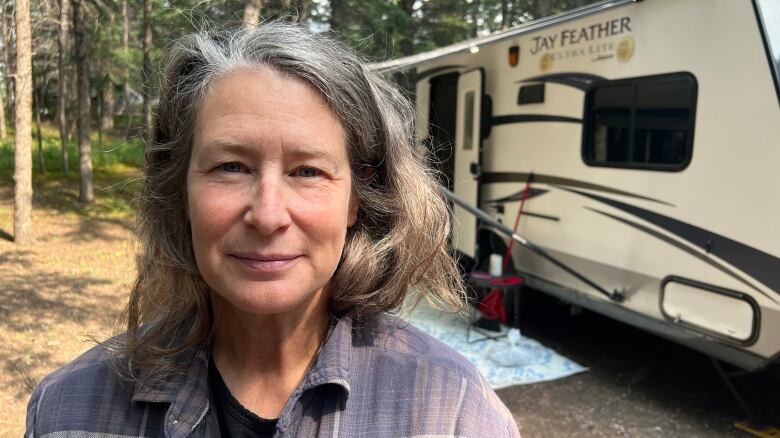 A white woman with long greying hair is wearing a lavender-coloured flannel shirt. She is standing outside, in a forested campground, in front of a camper trailer.