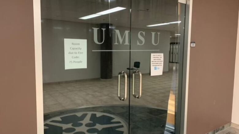 A glass door with the word 'UMSU' and some signs taped on it. there is a curtain behind the door.