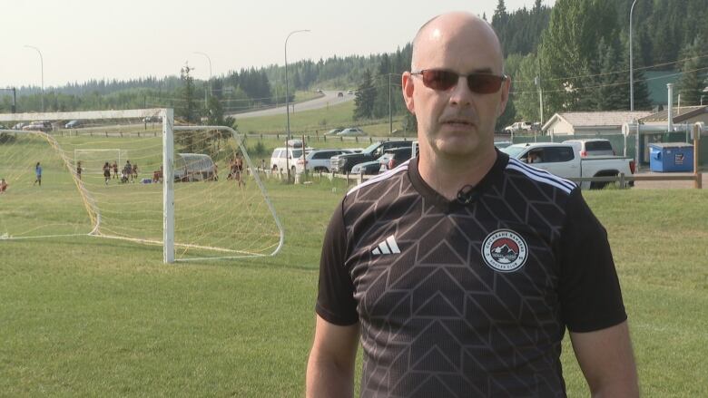 A bald man wearing black sunglasses is standing on a field talking into a camera.