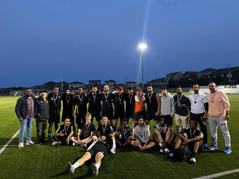 A group of men posing on the soccer field, most of them wearing soccer uniforms and medals around their necks.