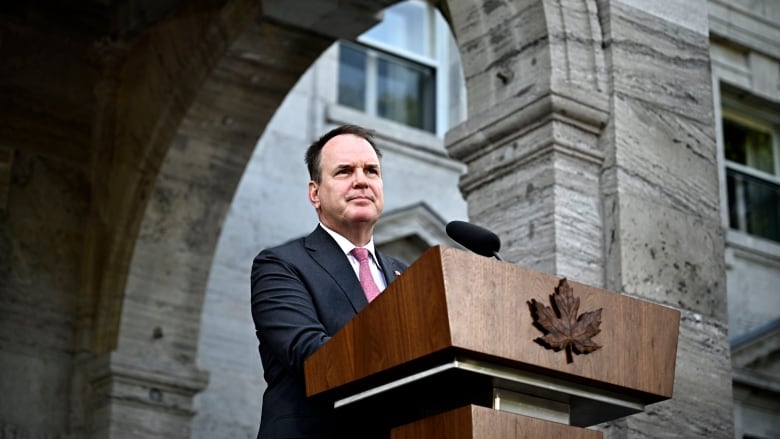 A man stands outside a stone edifice in front of a microphone attached to an expensive wood podium that is fronted by a wood carving of a maple leaf.