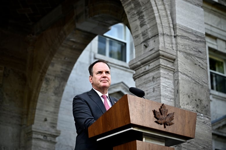 A man stands outside a stone edifice in front of a microphone attached to an expensive wood podium that is fronted by a wood carving of a maple leaf.