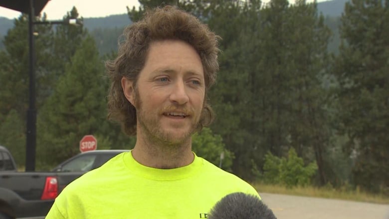 A man with a beard and bushy hair speaks in a parking lot.