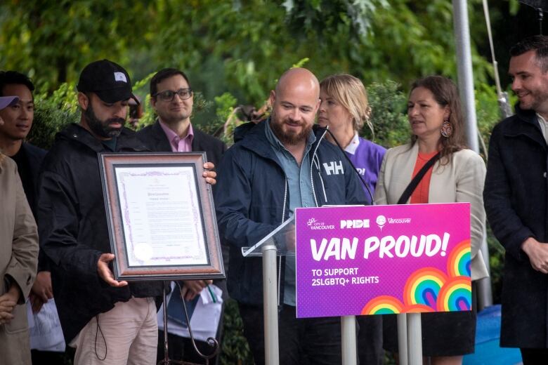 A bald man looks at a framed proclamation as he stands at a podium marked 'VAN PROUD! To support 2SLGBTQ+ rights'. He is surrounded by other people.