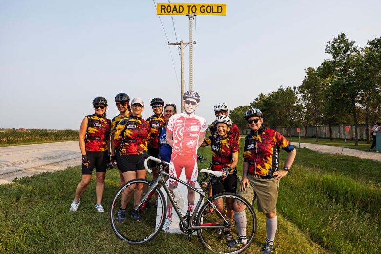 A group of people are standing around a cardboard cutout of a man wearing a cycling uniform, with a bike in front. A sign behind says 