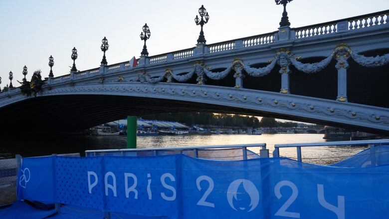 A bridge is shown spanning a river. Beneath it is a sign for the Paris 2024 Games.