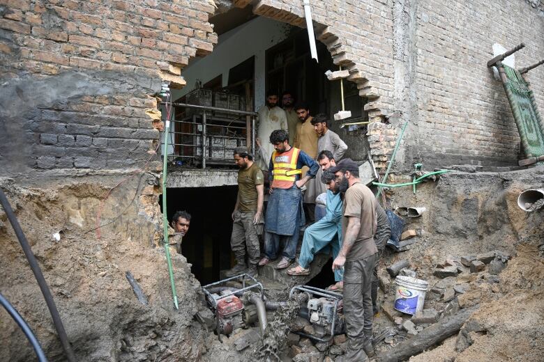 About seven or eight men are shown standing near a massive hole in the side of a building, with tools and debris seen on the dirt ground.