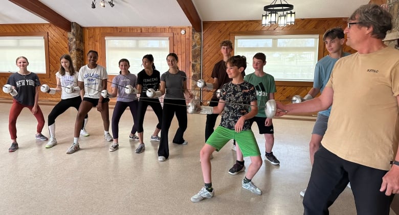 A group of kids, who are fencers, are seen holding their fencing blades as they pose for a picture with their coach, a man in a light shirt and black pants.