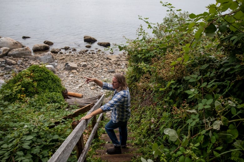 A man dressed casually in jeans and a checked shirt leans over a railing along a path near the ocean.