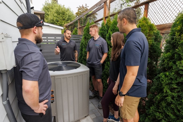 Contractor stands around heat pump with group of homeowners, explaining how a dual fuel system works.