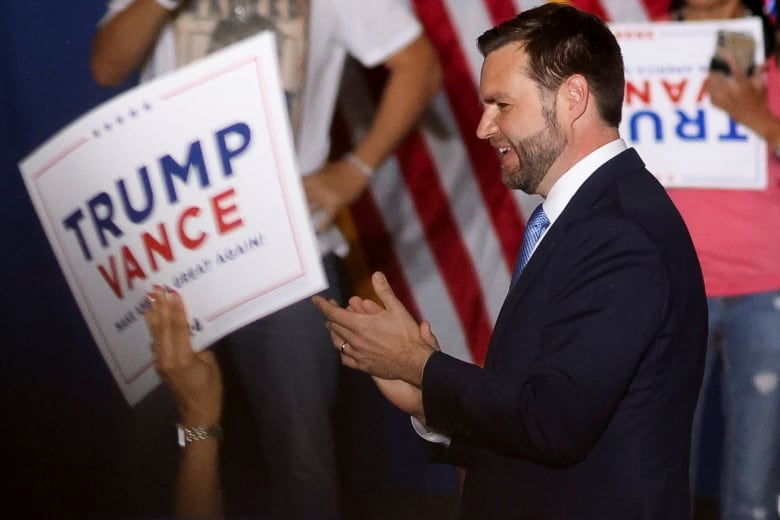 A bearded man in a suit, pictured in profile, smiles and claps in front of a crowd of people holding signs that read 'Trump/Vance.'